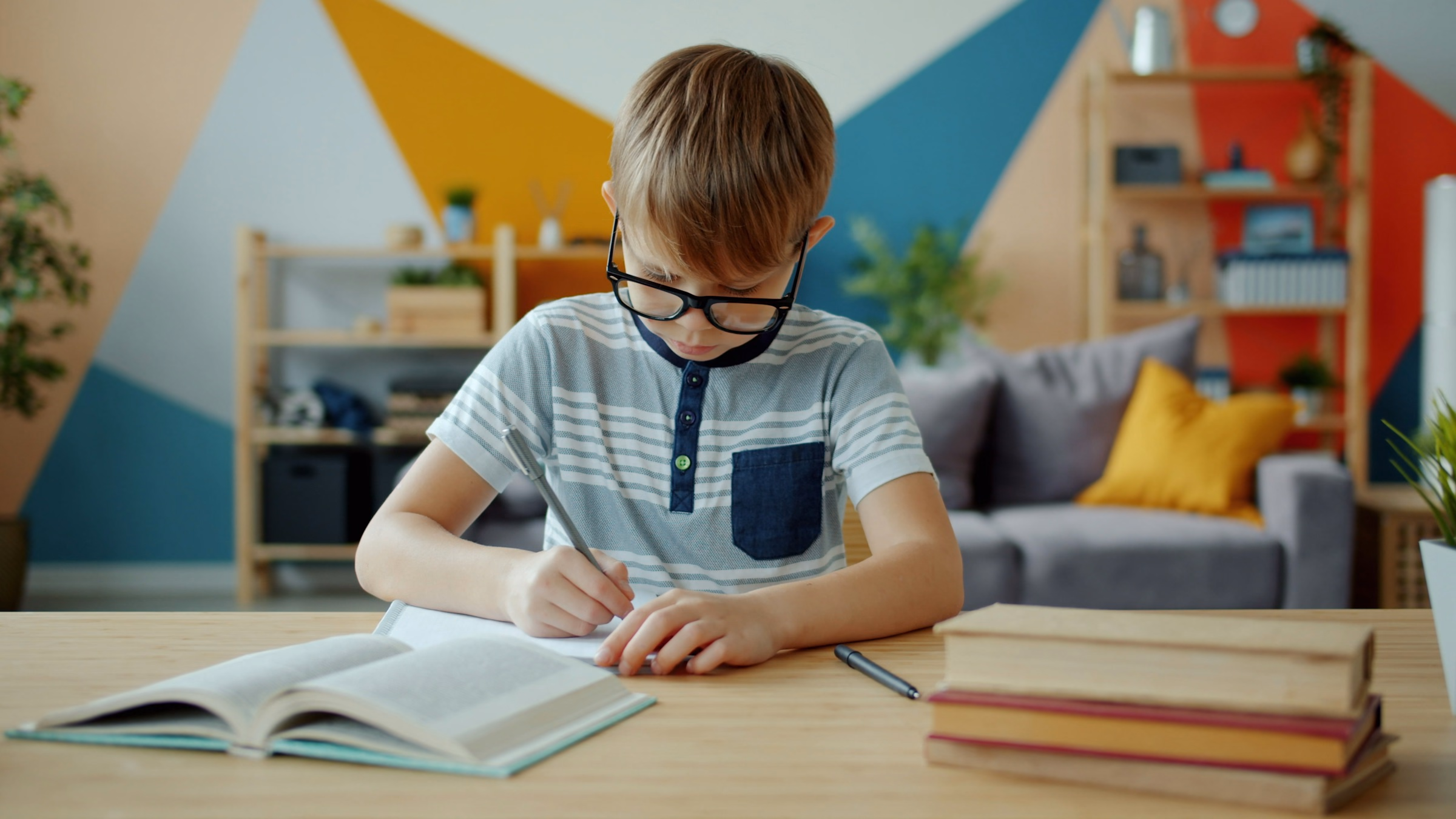 Boy in glasses writing in a notebook at a desk stacked with books