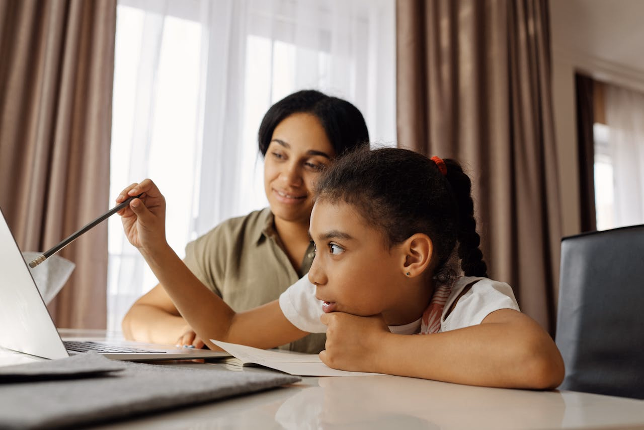 Girl pointing at a laptop screen with a pencil while her mother watches and smiles