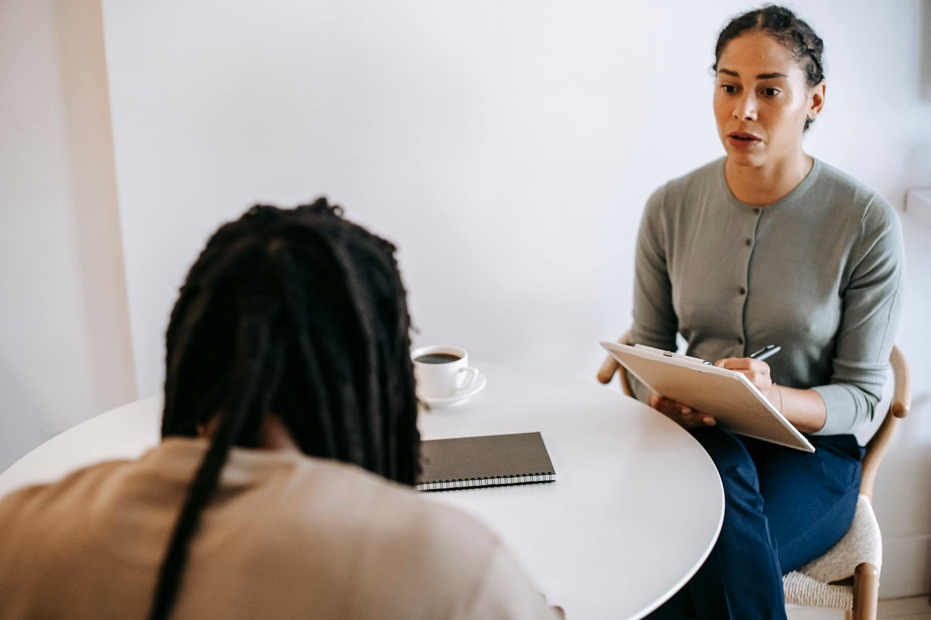 Clinician with a clipboard talking with a seated patient at a round white table