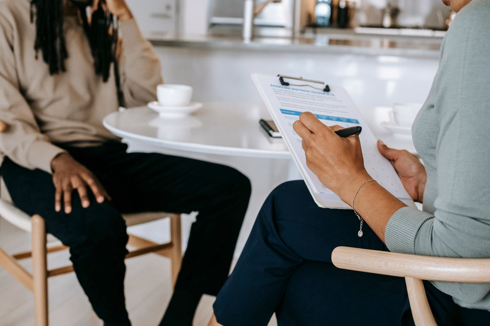Clinician taking notes on a clipboard while interviewing a patient across a white table