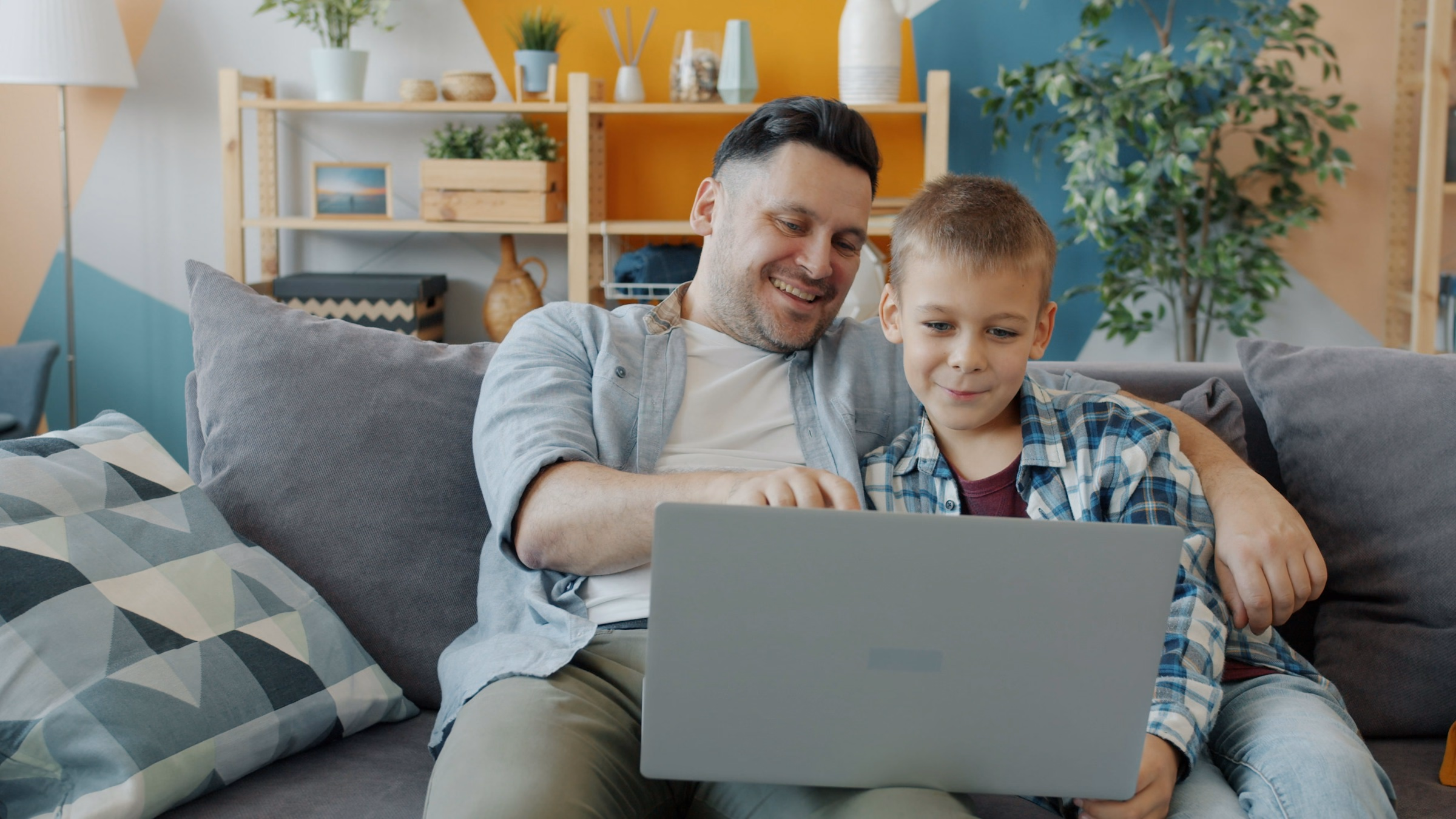 Father and son sitting close on a gray couch, looking at a laptop together