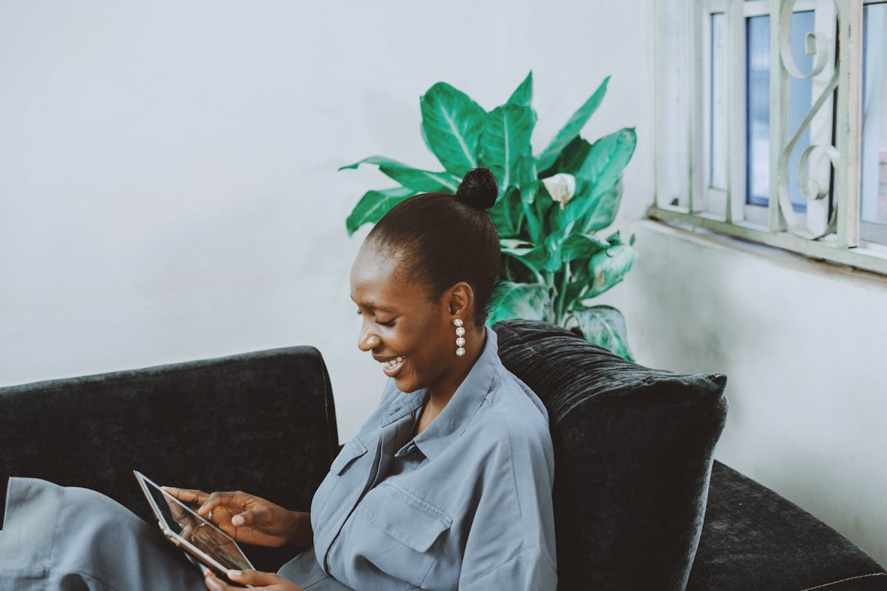 Woman on a dark couch joining a telepsychiatry visit on a tablet beside a leafy plant