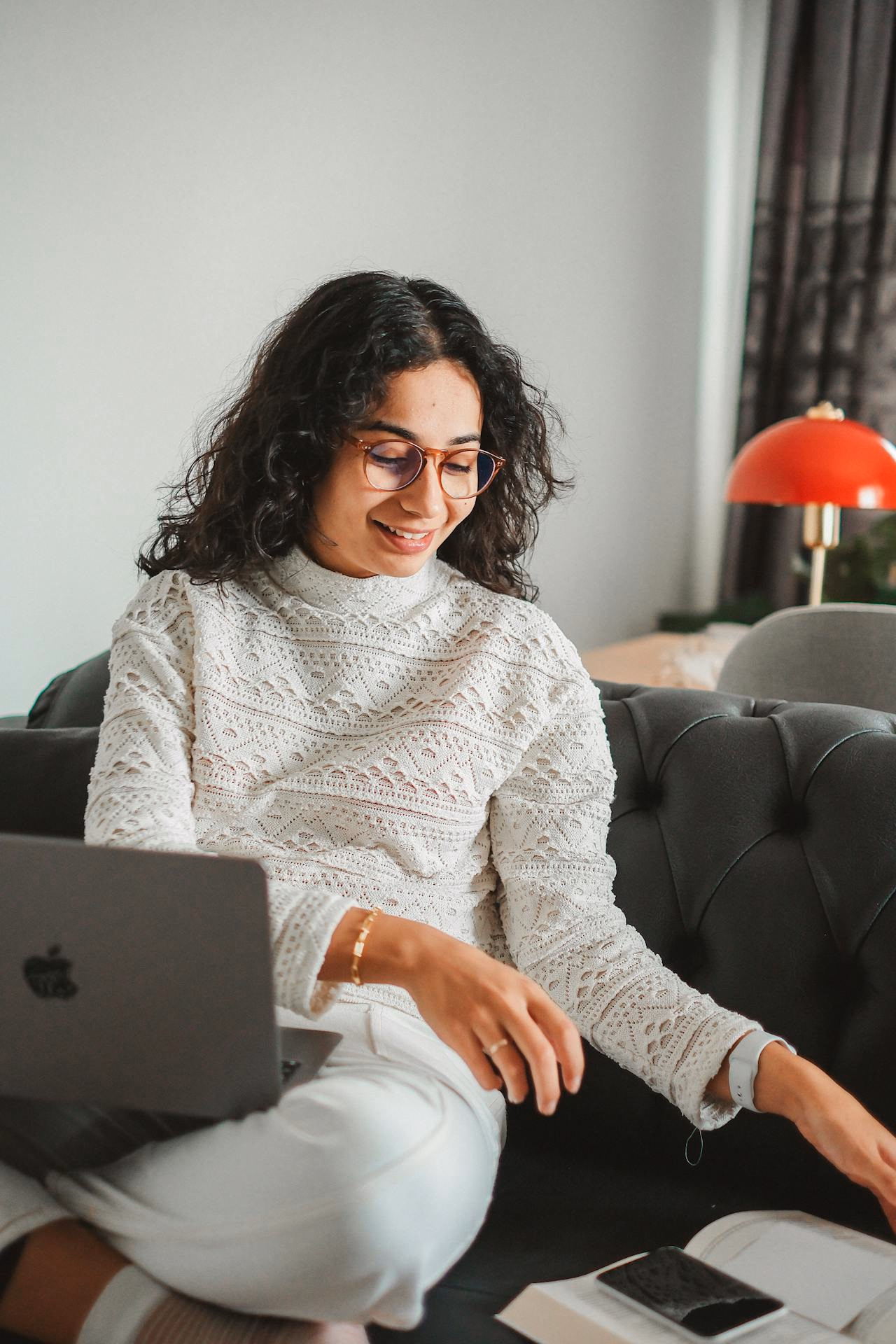 Woman in glasses on a couch with a laptop, reaching toward an open notebook and phone