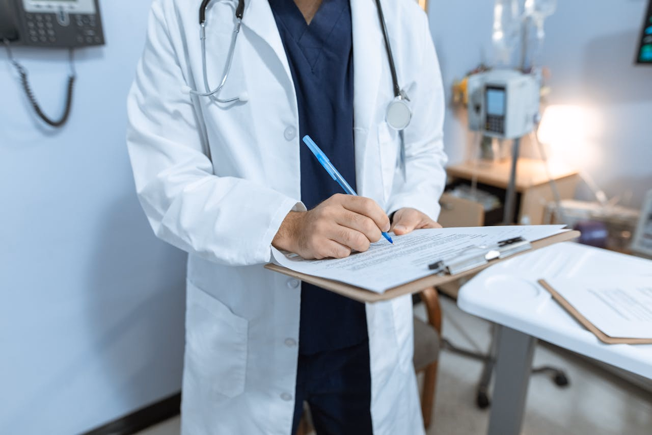 Prescriber in a white coat writing medication notes on a clipboard in a clinic office