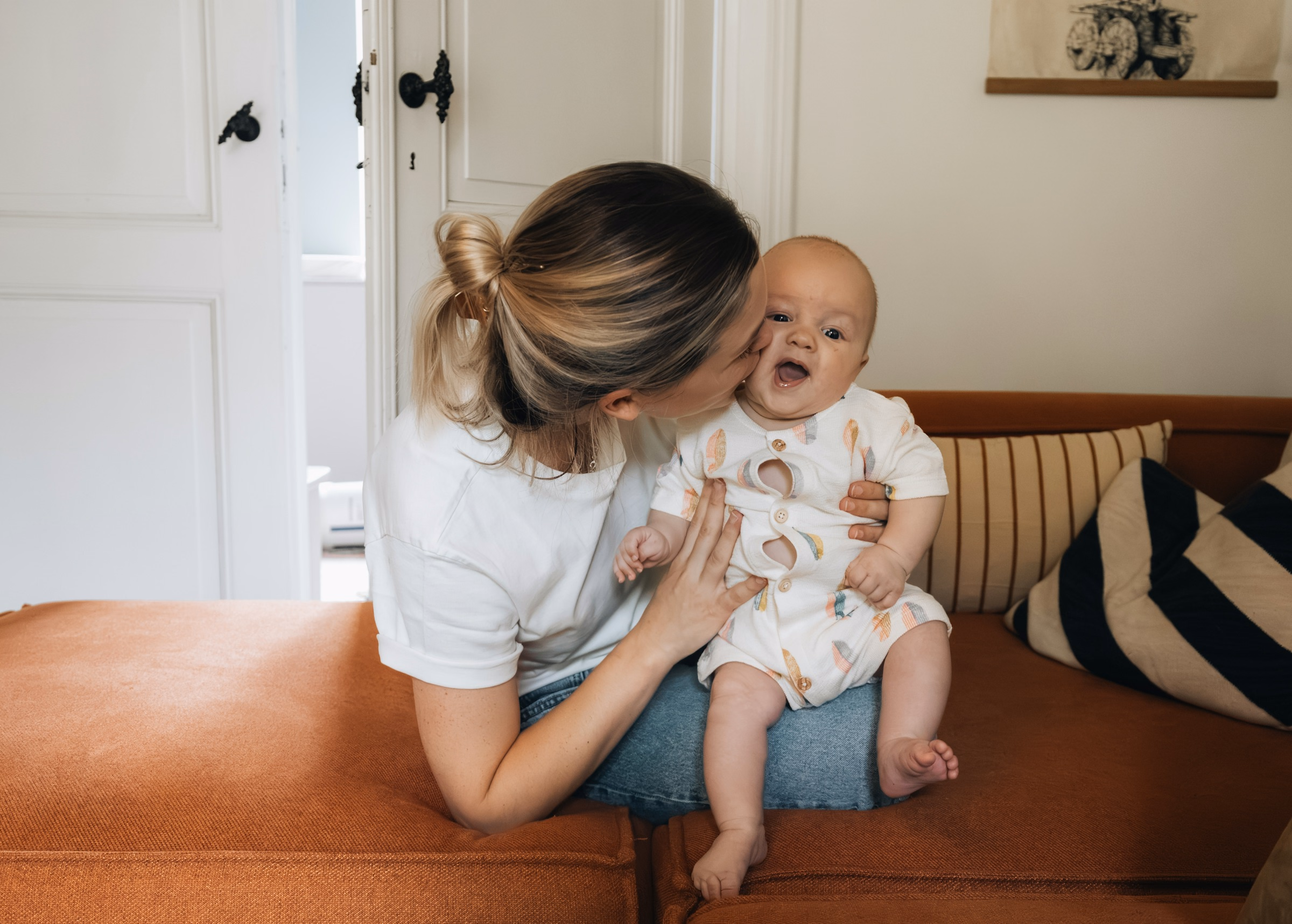 Mother kissing her smiling baby on an orange sofa at home