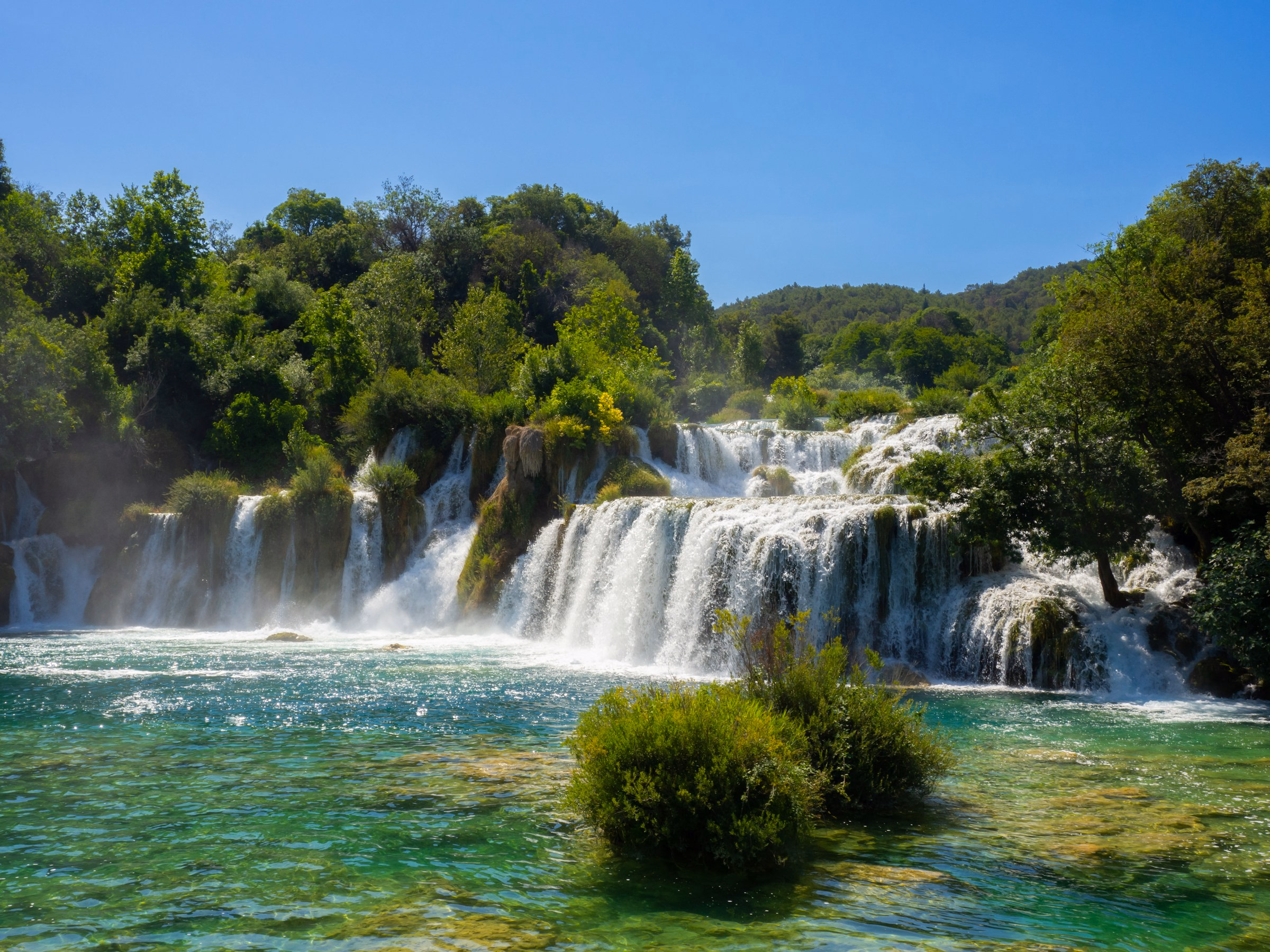 Waterfalls spilling into a clear green river below forested hills