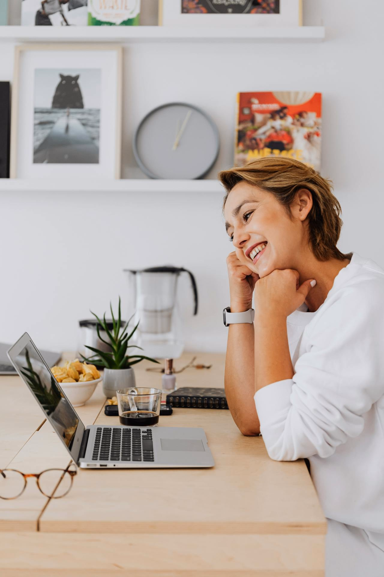 Woman at a wooden desk smiling at her laptop during a telepsychiatry appointment
