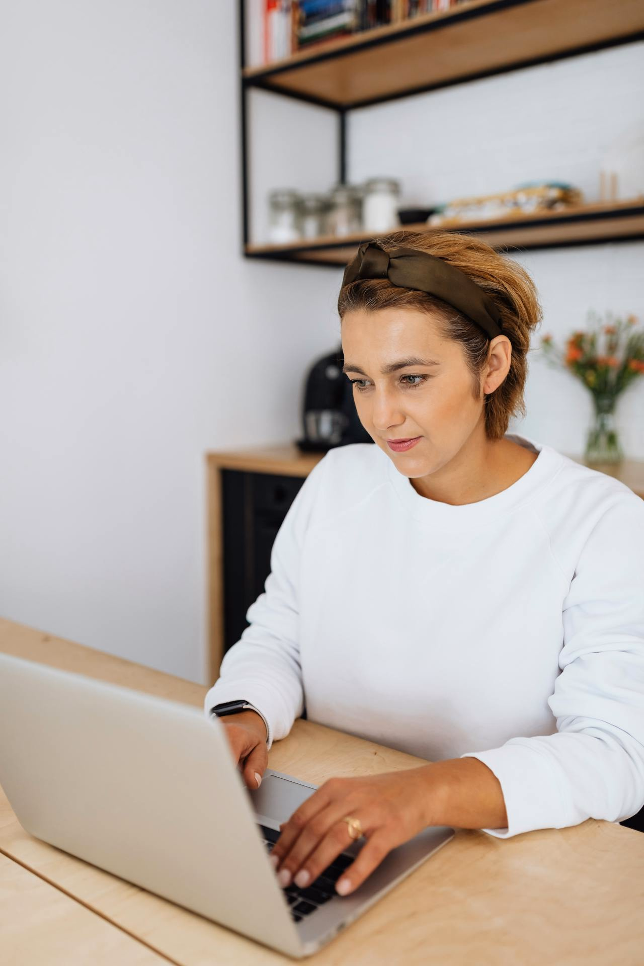 Woman in a white sweater and headband typing on a laptop at a kitchen desk