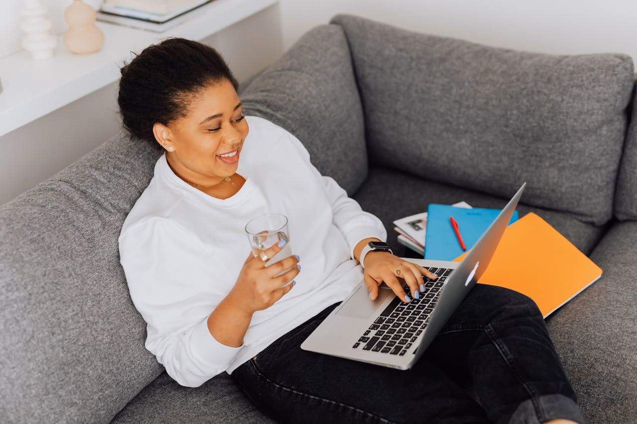Woman on a gray couch on a laptop video call, water glass in hand and folders beside her