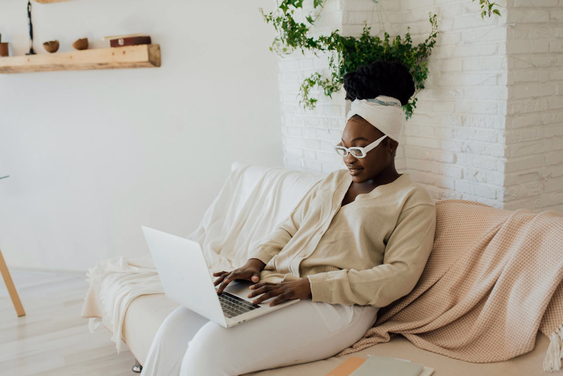 Woman in glasses and a headwrap on a sofa using a laptop among plants and light decor