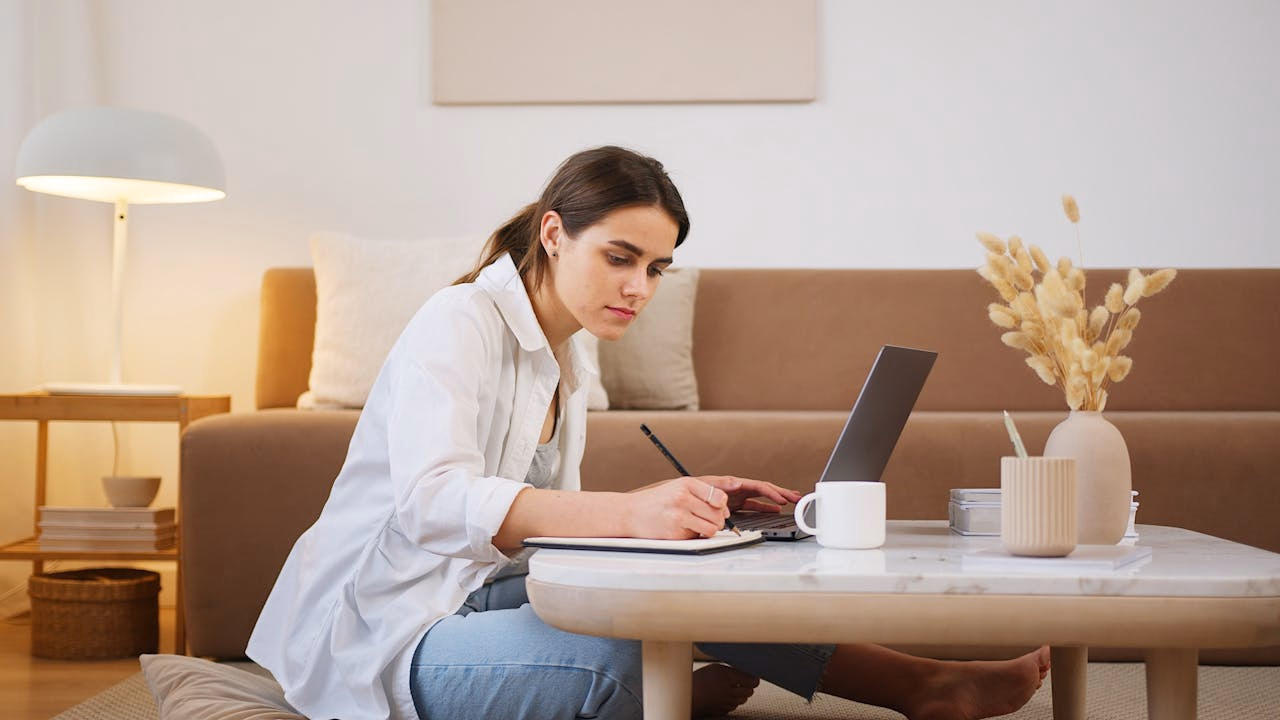 Woman writing in a notebook on the floor with a laptop open on the coffee table