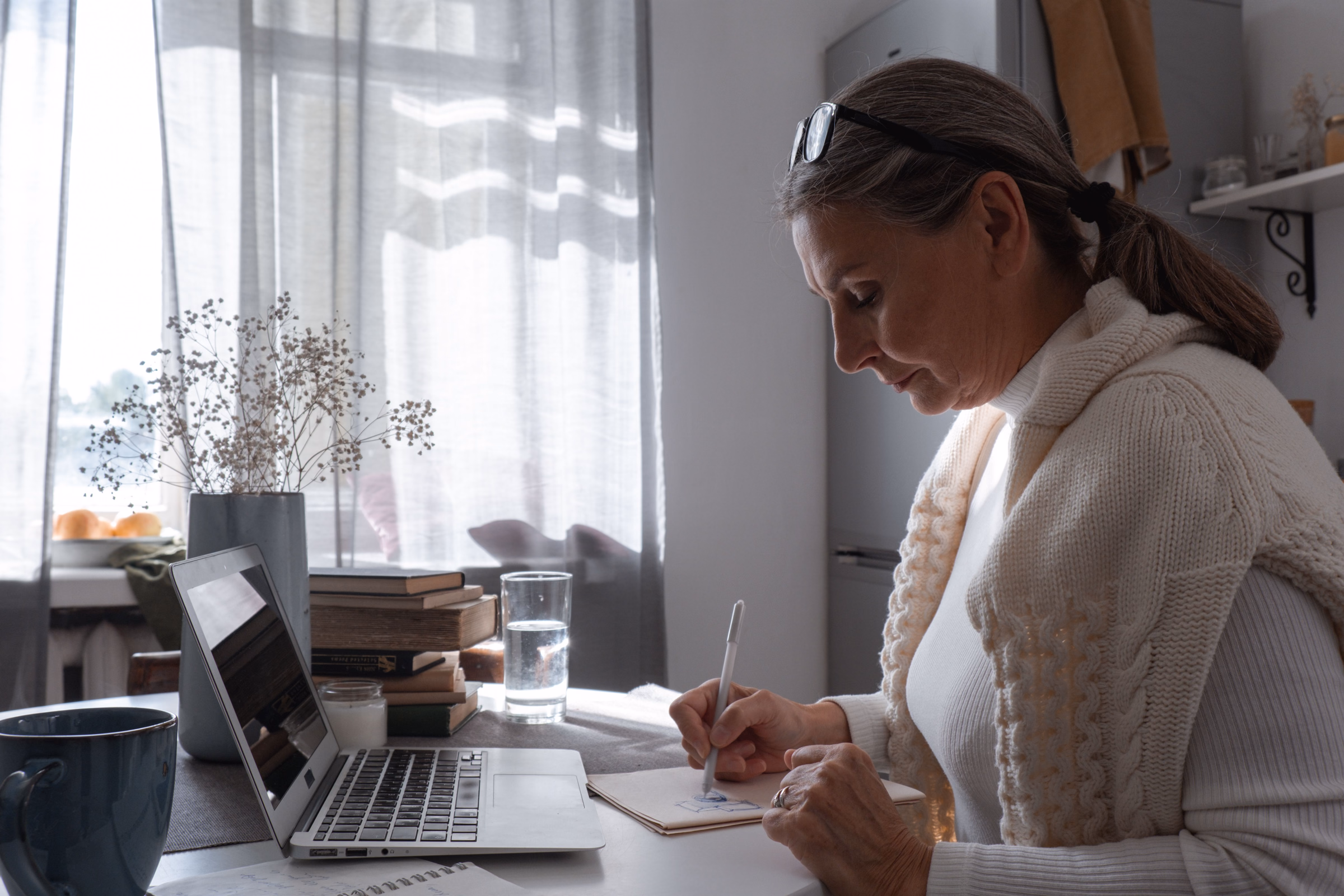 Woman in a cream sweater writing notes beside her laptop at home