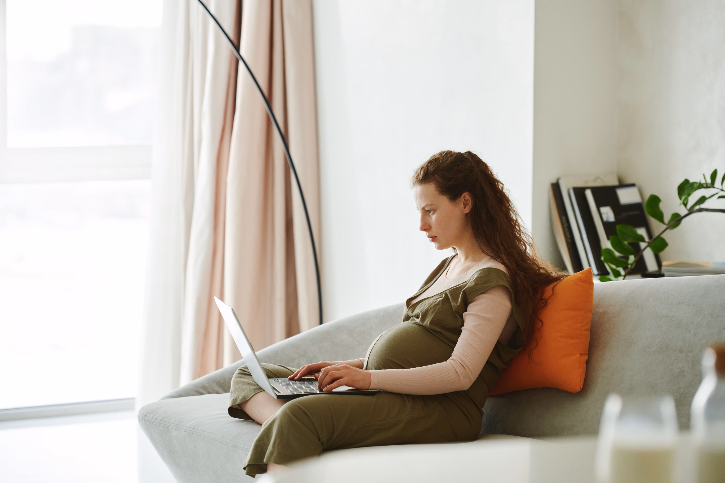 Pregnant woman on a gray sofa typing on a laptop in a bright living room
