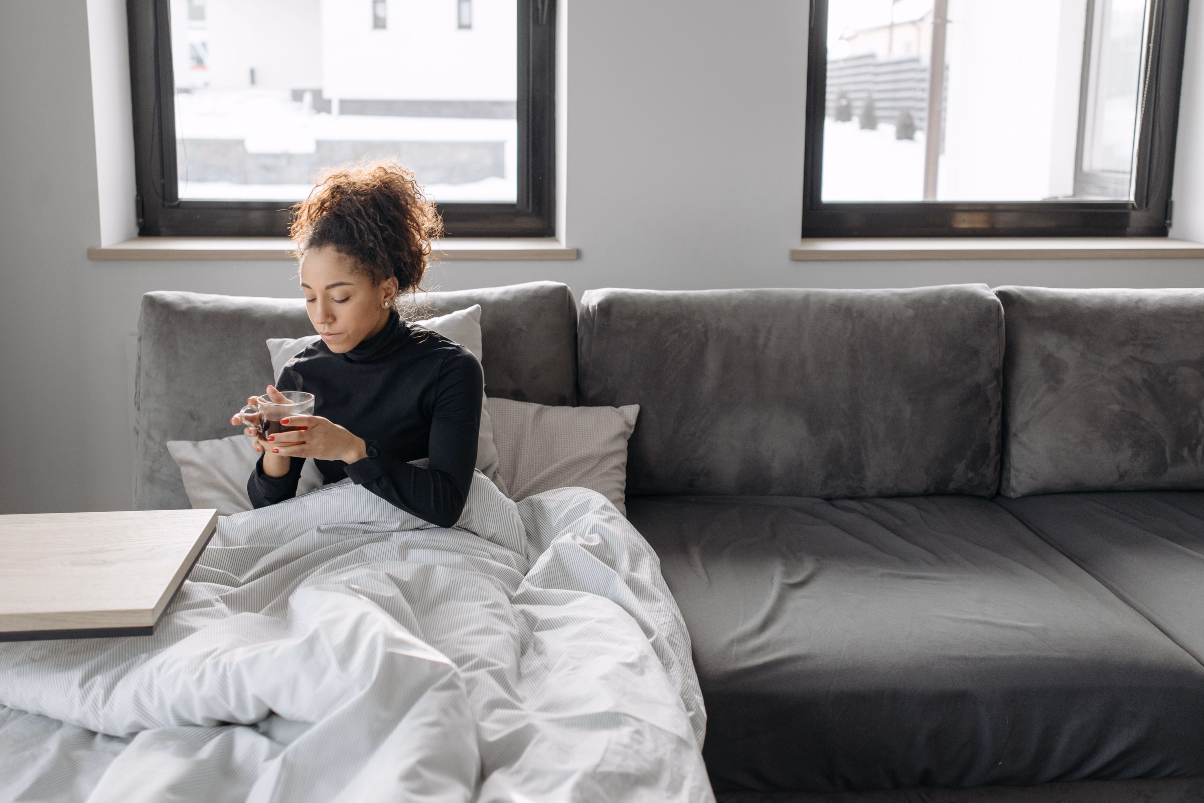 Woman wrapped in a white duvet on a gray sofa, holding a cup of tea