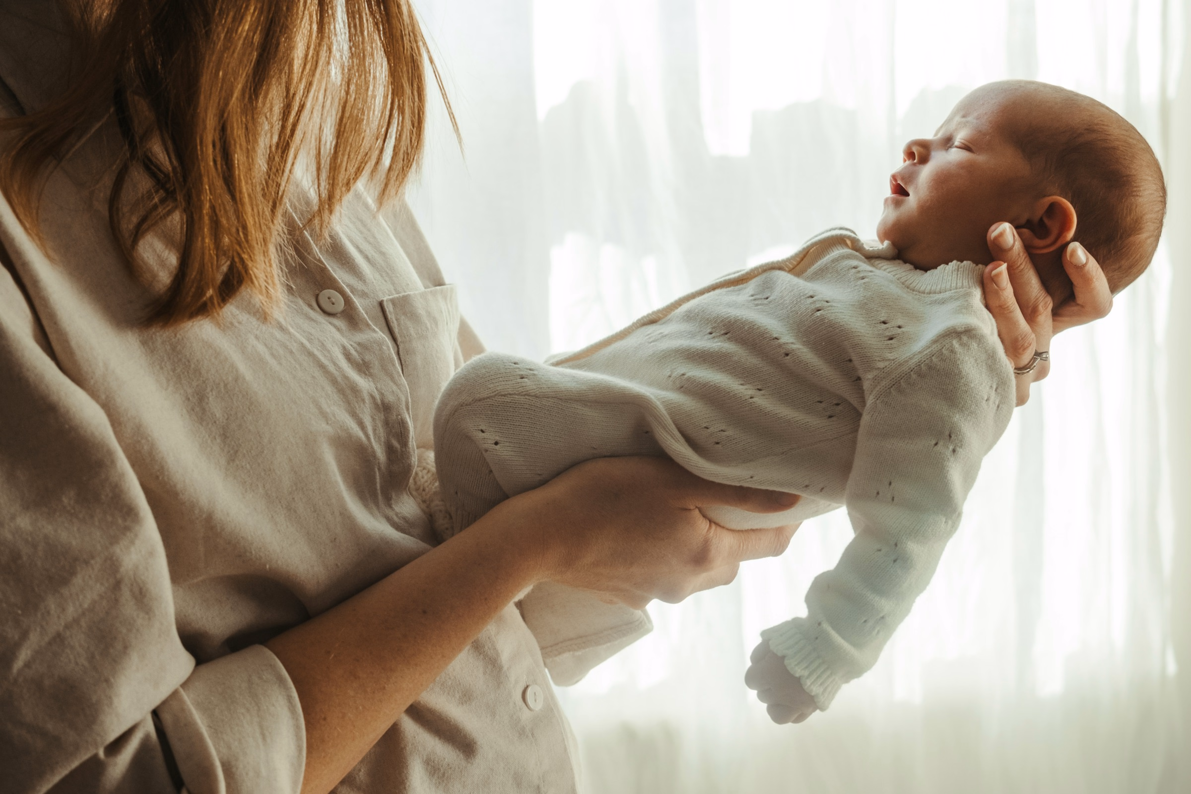Mother holding her sleeping newborn in front of a sheer curtained window