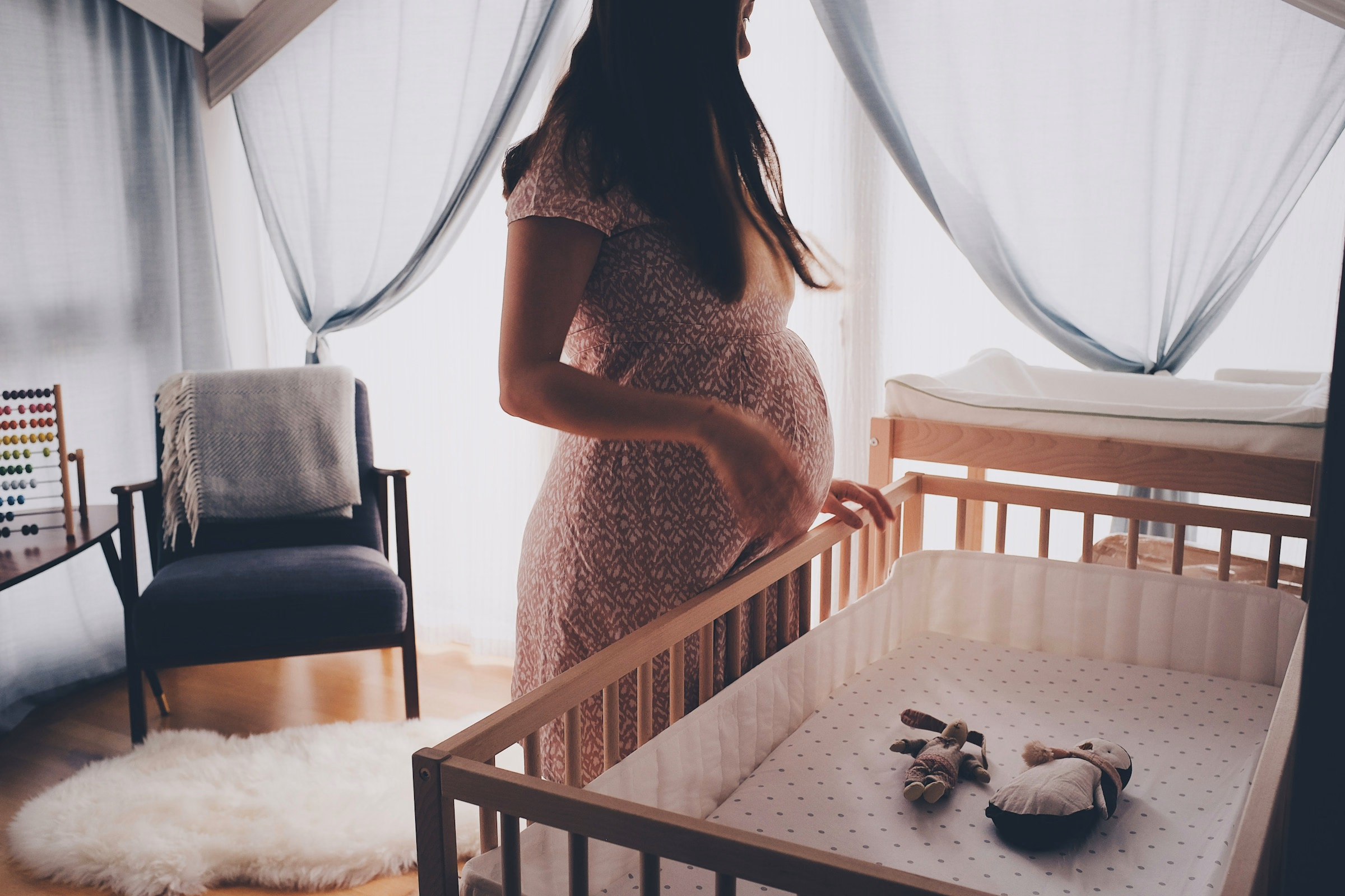 Pregnant woman resting a hand on a wooden crib in a sunlit nursery