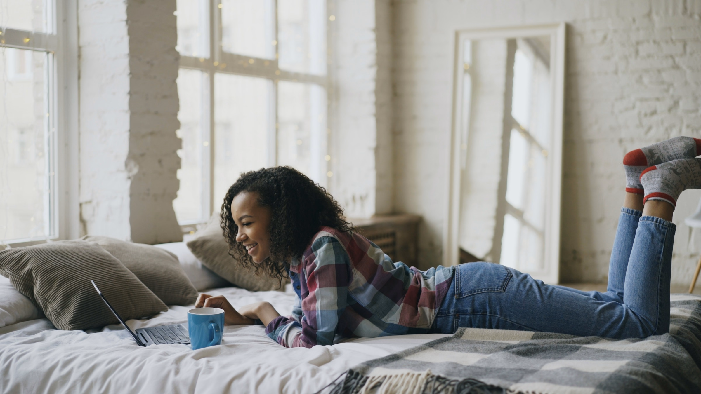 Teenage girl lying on her bed, smiling at a laptop beside a blue mug